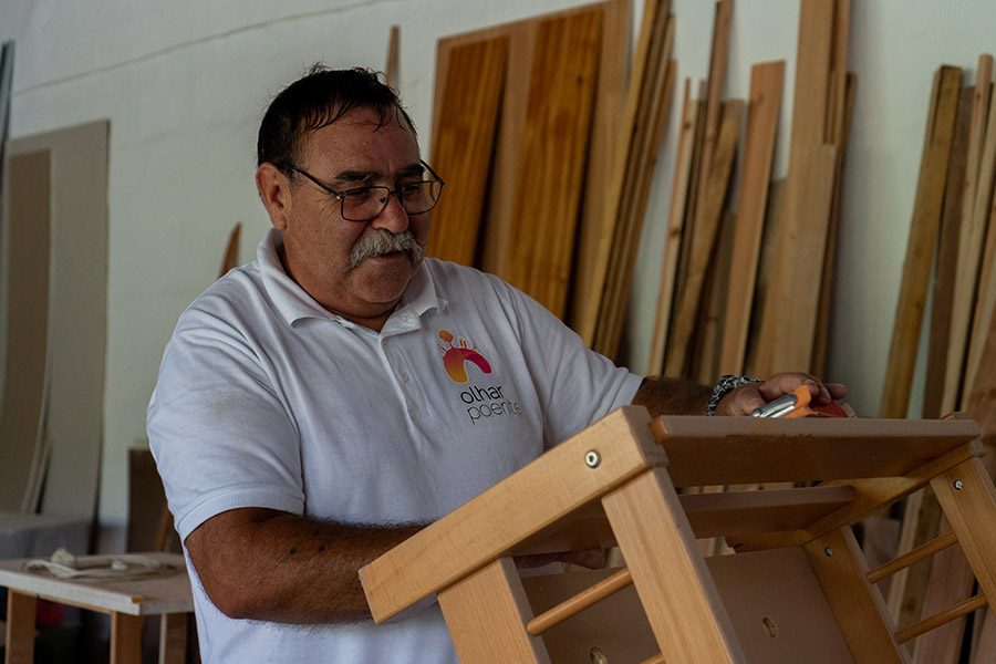 Técnico do SOS Casa, de pólo branco com o logótipo, trabalha numa peça de madeira numa oficina.
