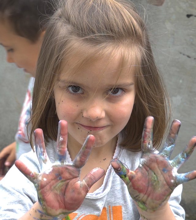 Menina sorridente mostra as mãos cobertas de tinta azul e verde.